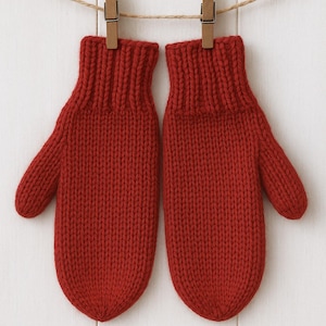 Op de afbeelding: Een paar gebreide rode wanten hangend aan een waslijn met houten knijpers. De wanten zijn gemaakt van dik, getextureerd garen en hebben een geribbelde manchet. De achtergrond is een wit houten oppervlak.