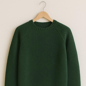 Può includere: Un maglione verde scuro a girocollo con maniche lunghe, appeso a una gruccia di legno. Il maglione ha polsini e orlo a coste. La maglia è di colore unito e sembra essere realizzata in un materiale morbido.