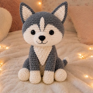 Puede incluir: Un peluche de ganchillo con forma de perro husky, hecho con hilo gris oscuro y blanco. El juguete tiene orejas puntiagudas con secciones interiores blancas, grandes ojos negros de plástico, nariz y boca bordadas en negro y patas blancas.