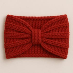 May include: A deep red knitted headband. The design features a central knot with ribbed detailing radiating outwards. The texture and colour suggest a warm accessory, ideal for colder weather. The headband is approximately 20 cm wide.