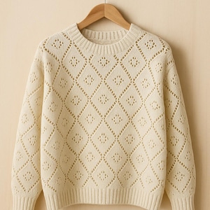 Könnte beinhalten: Cremefarbener Pullover mit einem Strickmuster in Rautenform. Der Pullover hat einen Rundhalsausschnitt und lange Ärmel. Das Strickmuster ist offen und erzeugt einen zarten, luftigen Look. Der Pullover hängt an einem Holzkleiderbügel.