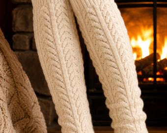 May include: A pair of cream-colored cable knit leg warmers featuring a vertical braided pattern and ribbed cuffs at the top and bottom. The leg warmers are made of a thick, textured knit fabric and extend from the mid-calf down to the ankle area.
