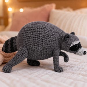 May include: A crocheted raccoon plush toy featuring a round dark gray body, a striped tail with alternating dark gray and black bands, and a face with a black eye mask, white muzzle, and small black nose, crafted with visible yarn stitches.