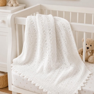 Peut inclure: Une couverture pour bébé tricotée en blanc avec un motif de dentelle à œillets délicat et une bordure festonnée. Le textile présente un design vertical répétitif de petits trous ajourés intégrés dans la structure du tissu tricoté.