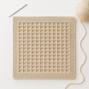 May include: A beige knitted square with a waffle-like texture, a border, and a needle. A ball of yarn is in the upper right corner. The square is likely a washcloth or a similar item.
