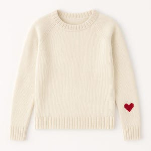 Pode incluir: Camisola creme com gola redonda e mangas compridas. Um coração vermelho é bordado na manga esquerda. A camisola tem punhos e bainha canelados. Malha de estilo clássico.