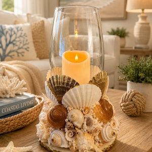 May include: A decorative candle holder featuring a large glass hurricane lamp. The base is adorned with a variety of seashells in shades of white, brown, and gold. A lit, ivory-colored candle sits inside, creating a warm glow. Coastal decor.