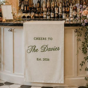 May include: A white fabric bar runner draped over a curved white bar counter. The runner features dark green text that reads "CHEERS TO The Davies Est. 2026". Large floral arrangements with pink, blue, and white flowers flank the bar runner.