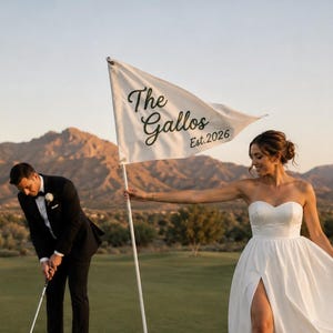 May include: A white triangular golf flag featuring the black text The Gallos Est. 2026 attached to a thin white pole. The flag is held by a person in a white strapless gown with a high leg slit, while another person in a black suit putts nearby.