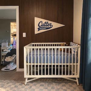 May include: A white wooden crib with a blue fitted sheet, two plush teddy bears, and a blue and white checkered pillow. Above the crib hangs a triangular cream-colored felt pennant featuring the blue text Cutter and the name Michael below it.