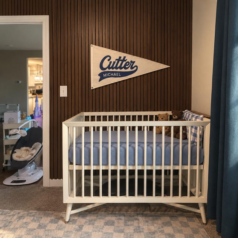 May include: A white wooden crib with a blue fitted sheet, two plush teddy bears, and a blue and white checkered pillow. Above the crib hangs a triangular cream-colored felt pennant featuring the blue text Cutter and the name Michael below it.