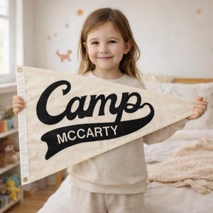 May include: A triangular off-white canvas pennant flag featuring the word Camp in large black script lettering above the word MCCARTY in a bold black banner graphic. The pennant includes a white side strip with two gold-toned metal grommets.