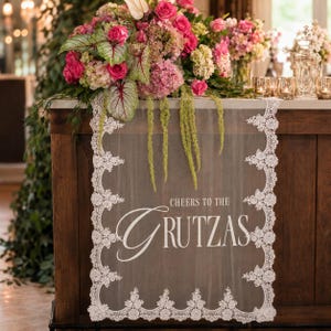 May include: A white lace table runner featuring a scalloped edge with floral embroidery and the printed text "CHEERS TO THE GRUTZAS" in the center. The runner is draped over a wooden surface beneath a large arrangement of pink and green flowers.