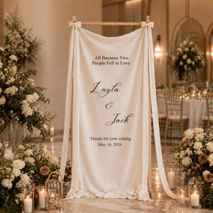 May include: A cream-colored wedding sign with the text "All Because Two People Fell in Love," the names "Layla & Jack," and the date "May 16, 2026." The sign is draped on a wooden frame, with floral arrangements and candles in the background.