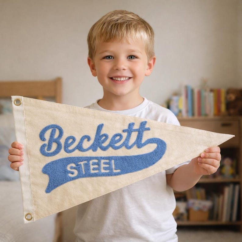 May include: A triangular off-white felt pennant featuring the name Beckett in a large blue script font above the word STEEL in a smaller blue block font. The pennant has two metal grommets on the left side for hanging and a textured finish.