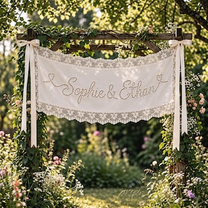 May include: A rectangular off-white fabric banner featuring the embroidered names Sophie & Ethan in a cursive script. The banner is bordered by intricate white lace trim and is suspended by two long, smooth cream-coloured satin ribbons.