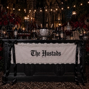 以下が含まれることがあります： 黒いバーテーブルに、黒いゴシック体で「The Hustads」と書かれた白いレースのランナーが掛けられています。テーブルの上には、大きな銀のボウル、複数のワインボトル、グラス、そして濃い赤色のフラワーアレンジメントが飾られています。