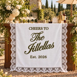 以下が含まれることがあります： 「CHEERS TO The Jillellas Est. 2026」というダークグリーンの文字がプリントされた白い長方形のテーブルランナー。ランナーの四辺は、花柄の複雑で幅広な白いレースのトリムで縁取られており、装飾的な仕上がりになっています。