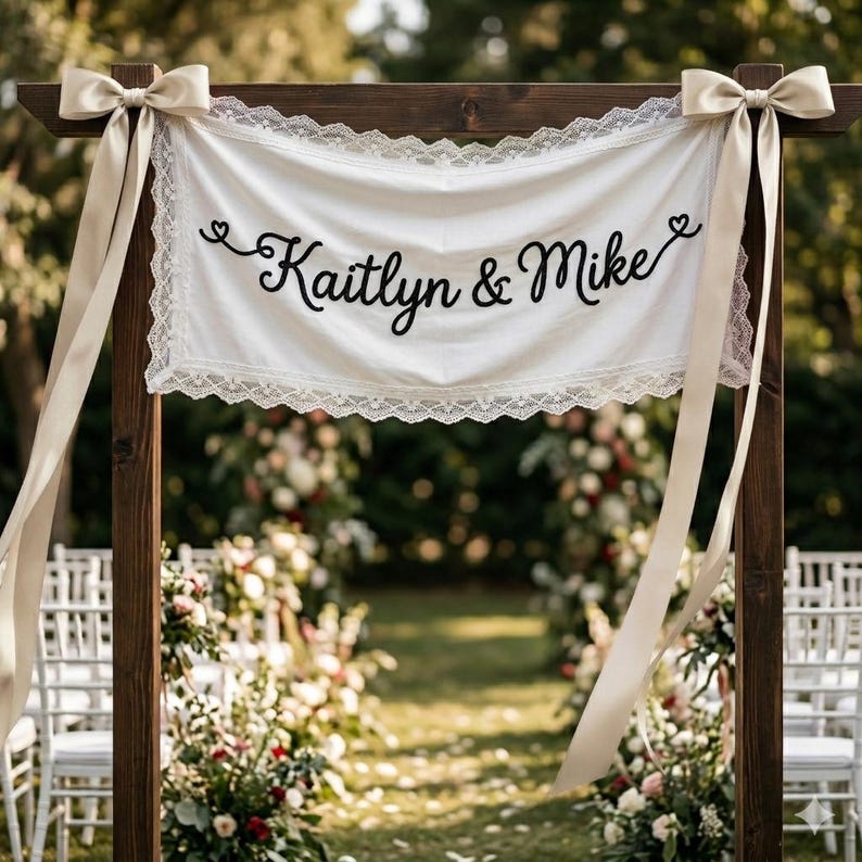 K&ouml;nnte beinhalten: Ein wei&szlig;es Stoffbanner mit Spitzenbesatz und dem schwarzen Schriftzug "Kaitlyn & Mike" in Schreibschrift. Das Banner h&auml;ngt an einem dunklen Holzrahmen, der an beiden Seiten des oberen Querbalkens mit gro&szlig;en beigen Satinschleifen verziert ist.
