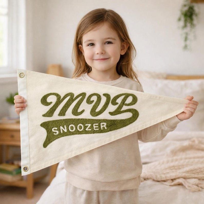 May include: A triangular off-white felt pennant flag featuring the text MVP SNOOZER in raised, textured olive green lettering. The pennant includes a white vertical side strip with two brass-colored metal grommets for hanging purposes.
