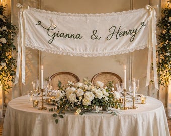 May include: A cream-colored fabric banner featuring the embroidered text "Gianna & Henry" in dark green script, accented with lace trim and tied with ribbons at both ends. The banner is displayed above a round table with a floral centerpiece.