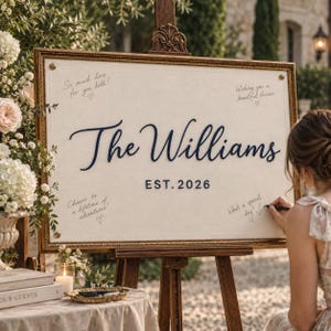 May include: A rectangular sign in a gold frame on a wooden easel featuring the text The Williams and EST. 2026 in dark blue script and block lettering. The off-white fabric surface includes handwritten messages and small heart illustrations.