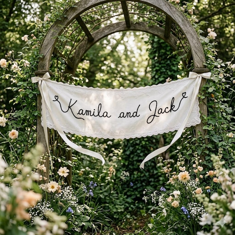 K&ouml;nnte beinhalten: Ein wei&szlig;es Stoffbanner mit gewelltem Spitzenrand, das den in Schwarz gestickten Text "Kamila and Jack" sowie zwei kleine Herzsymbole zeigt. Das Banner ist mit wei&szlig;en B&auml;ndern an einem h&ouml;lzernen Gartenbogen befestigt.