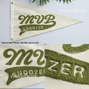 May include: A triangular off-white canvas pennant featuring the embroidered olive green text MVP and SNOOZER. The pennant includes a white vertical strip with two metal grommets on the left side for hanging, showcasing detailed stitching.