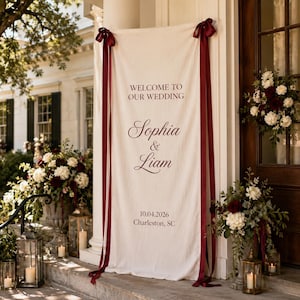 May include: A vertical off-white fabric wedding banner featuring dark red text that reads WELCOME TO OUR WEDDING, Sophia & Liam, 10.04.2026, and Charleston, SC. The banner is secured at the top corners with long, dark red satin ribbons.