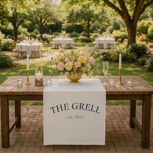 Könnte beinhalten: Ein Holztisch für eine Outdoor-Veranstaltung, mit einem weißen Tischläufer mit dem Aufdruck "THE GRELL est. 2025". Der Tisch ist mit einem Blumenarrangement, Kerzen und Sektgläsern dekoriert. Im Hintergrund sind weitere Tische und Bäume zu sehen.