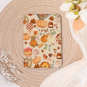 May include: A beige notebook featuring a fall-themed pattern. The design includes pumpkins, leaves, a teddy bear, and other autumn-related illustrations. The notebook is rectangular and likely used for writing or sketching.