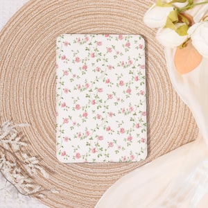 Peut inclure: Un carnet rectangulaire à motifs floraux, avec un fond blanc orné de roses roses et de feuilles vertes. Le carnet est posé sur un fond beige tissé, avec un tissu crème et des fleurs dans le coin supérieur droit.