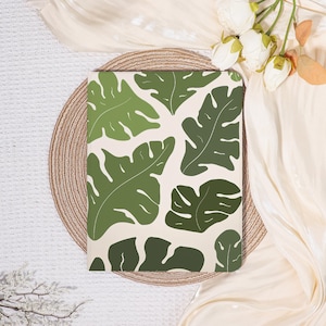 May include: A notebook with a cream-colored background and a pattern of green monstera leaves. The notebook is placed on a woven placemat and surrounded by white flowers and fabric.