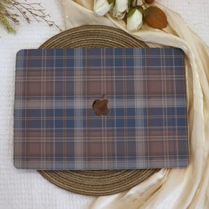 Könnte beinhalten: Eine Laptop-Hülle mit blauem und braunem Karomuster und einem Apple-Logo-Ausschnitt. Die Hülle hat ein klassisches Tartanmuster in Blau-, Braun- und Beigetönen. Die Laptop-Hülle ist für ein MacBook konzipiert.