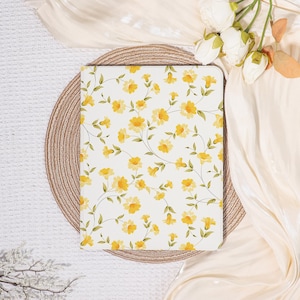 May include: A rectangular notebook with a white background and a yellow floral pattern. The flowers are in full bloom and have green stems and leaves. The notebook is resting on a round, woven placemat.