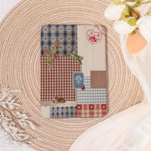 May include: A decorative notebook with a patchwork design. The cover features various patterns, including stars, gingham, and cable knit, in shades of blue, red, and cream. A small floral arrangement and a green ribbon add detail. A heart-shaped embroidery is also visible.