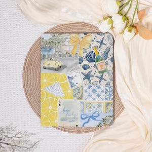 May include: A decorative notebook with a collage of Italian-themed illustrations. The design includes a yellow car, lemons, seashells, and the text "how lucky are we". The notebook has a yellow bow and a champagne bottle illustration.