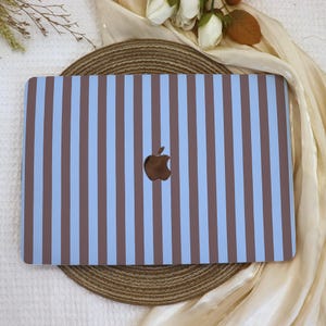 May include: A laptop skin featuring a vertical striped pattern in alternating light blue and dark brown colors. The rectangular cover has a centered cutout revealing the metallic Apple logo, maintaining a clean and symmetrical aesthetic across the surface.