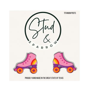 May include: A pair of pink roller skate earrings with purple bases and orange wheels, featuring a rainbow graphic on the side. The display card includes the text Stud & Sparrow, TITANIUM POSTS, and PROUDLY HAND MADE IN THE GREAT STATE OF TEXAS.