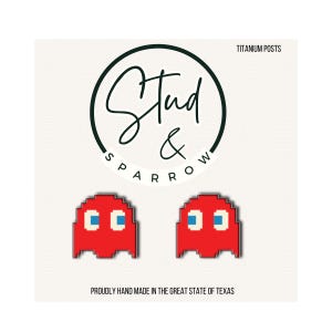 May include: A pair of red pixelated ghost earrings with blue and white details. The earrings are in the shape of a classic video game character. The text "Stud & Sparrow" is in a circle above the earrings. The text "TITANIUM POSTS" is above the circle.