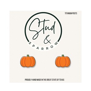 May include: A pair of orange pumpkin-shaped stud earrings. The earrings are set against a white background with the text "Stud & Sparrow" in a circular design. The text "Titanium Posts" and "Proudly Hand Made in the Great State of Texas" are also visible.