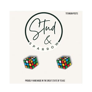 May include: A pair of colorful Rubik's Cube stud earrings. The earrings are square-shaped with a classic multi-colored pattern. The image includes the text "Stud & Sparrow" and "Titanium Posts". The earrings are proudly hand made in the Great State of Texas.