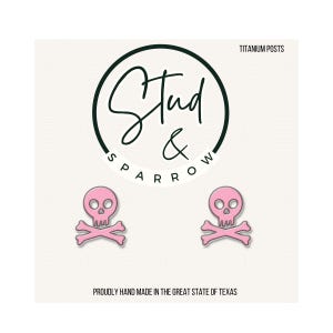 May include: Pink skull and crossbones earrings. The earrings are made of pink enamel and have a skull and crossbones design. The text "Stud & Sparrow" is in a black circle. The text "TITANIUM POSTS" is at the top. The text "PROUDLY HAND MADE IN THE GREAT STATE OF TEXAS" is at the bottom.