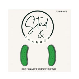 May include: Green pickle-shaped stud earrings. The earrings are on a white background with the text "Stud & Sparrow" in a circular design. The text "Titanium Posts" is above the earrings, and "Proudly hand made in the great state of Texas" is below.