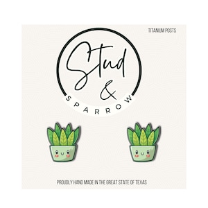 May include: A pair of green and yellow succulent plant stud earrings with smiling faces in light green pots. The earrings are displayed on a white background with the text "Stud & Sparrow" and "Titanium Posts". The text "Proudly hand made in the great state of Texas" is also visible.