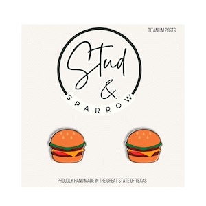 May include: A pair of hamburger-shaped stud earrings with titanium posts, displayed on a white card featuring the Stud & Sparrow logo and the text PROUDLY HAND MADE IN THE GREAT STATE OF TEXAS. The burgers feature orange buns, green lettuce, and yellow cheese.