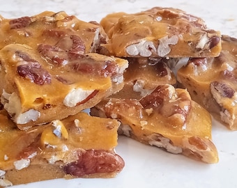 May include: A close-up of a plate of pecan brittle, a sweet and crunchy treat made with pecans, sugar, and butter. The brittle is broken into irregular pieces and has a golden brown color.