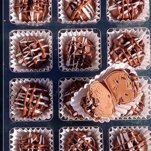 Pu&ograve; includere: Una scatola di tartufi al cioccolato assortiti con cioccolato bianco e marrone glassato sopra. I tartufi sono disposti in una griglia in una scatola di plastica trasparente.