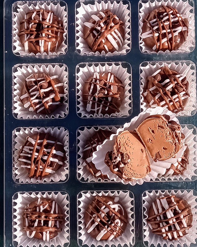 Pu&ograve; includere: Una scatola di tartufi al cioccolato assortiti con cioccolato bianco e marrone glassato sopra. I tartufi sono disposti in una griglia in una scatola di plastica trasparente.