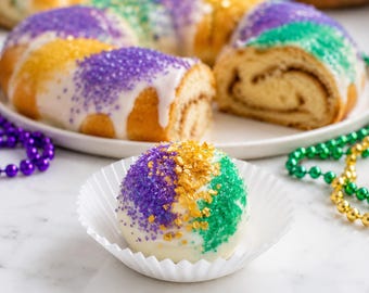 Può includere: Un cake pop rotondo ricoperto di glassa bianca e guarnito con cristalli di zucchero viola, oro e verdi, adagiato in un pirottino di carta bianco. Sullo sfondo, un dolce a forma di ciambella tagliato presenta decorazioni simili.
